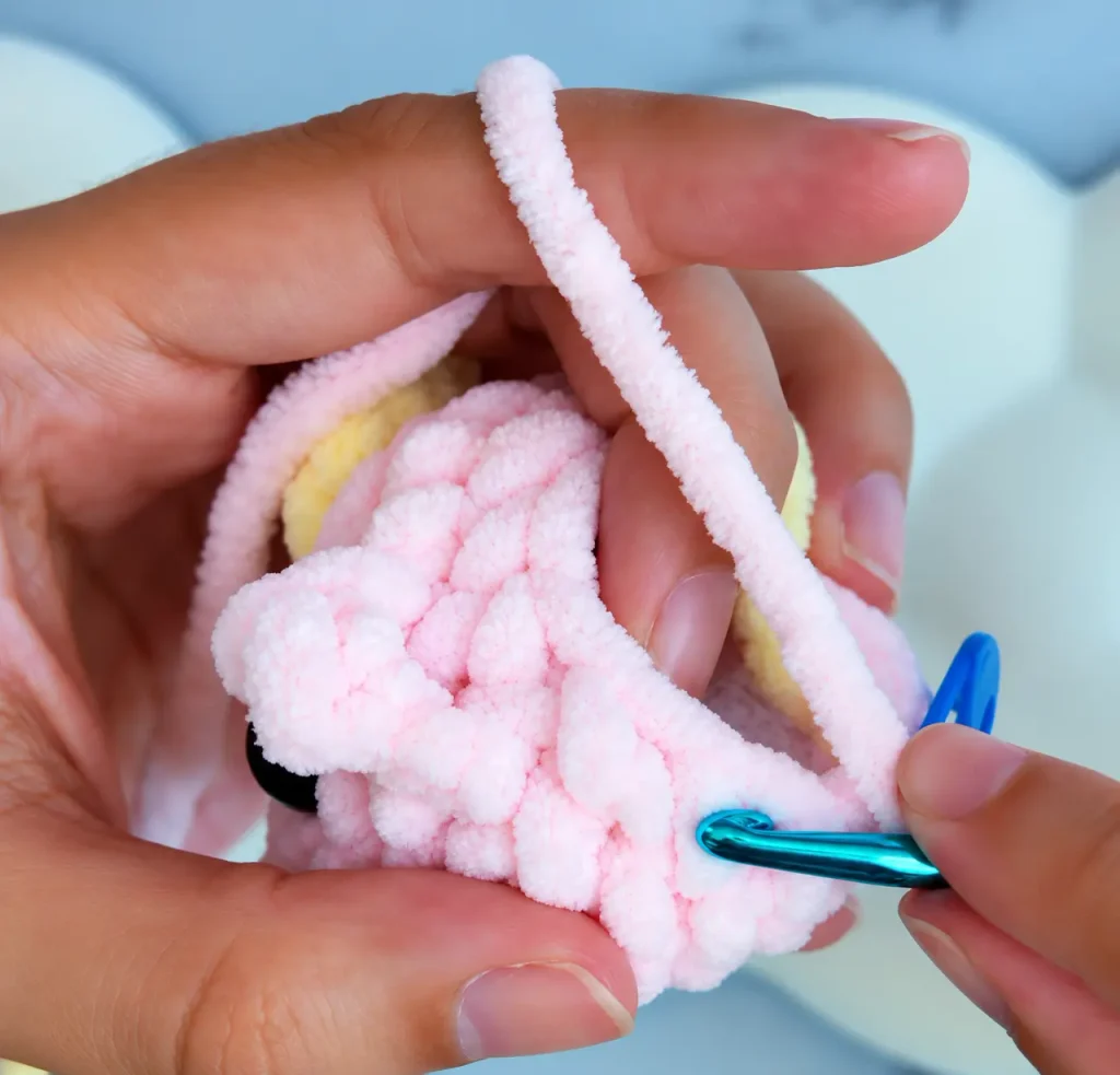 Close-up of a blue crochet hook working pink chenille stitches while shaping the dragon's head in Round 8
